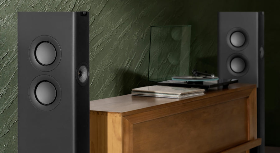 Two modern floor-standing speakers positioned on either side of a wooden cabinet with a turntable and vinyl records, set against a textured dark green wall.