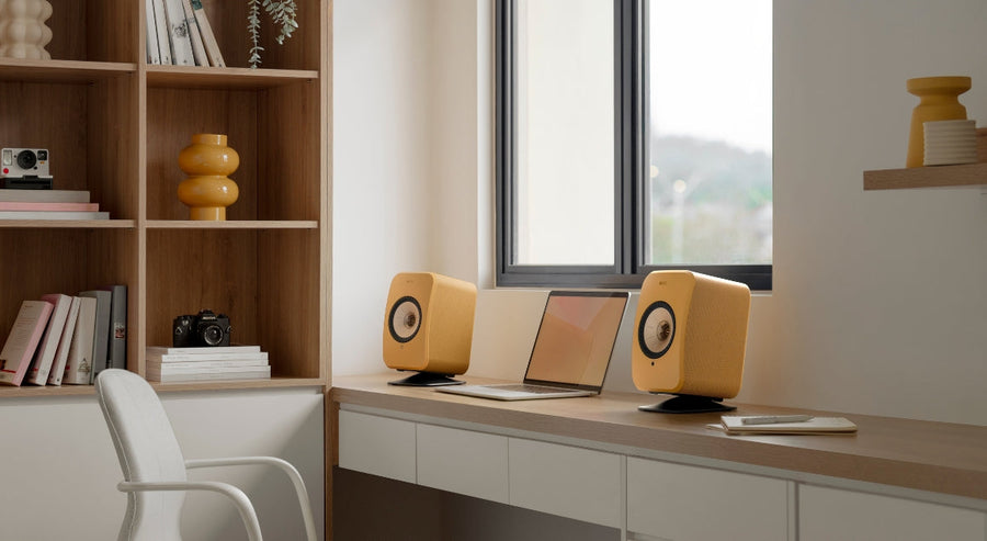 KEF wireless bookshelf speakers in yellow on a desk with a laptop in a modern home office setup