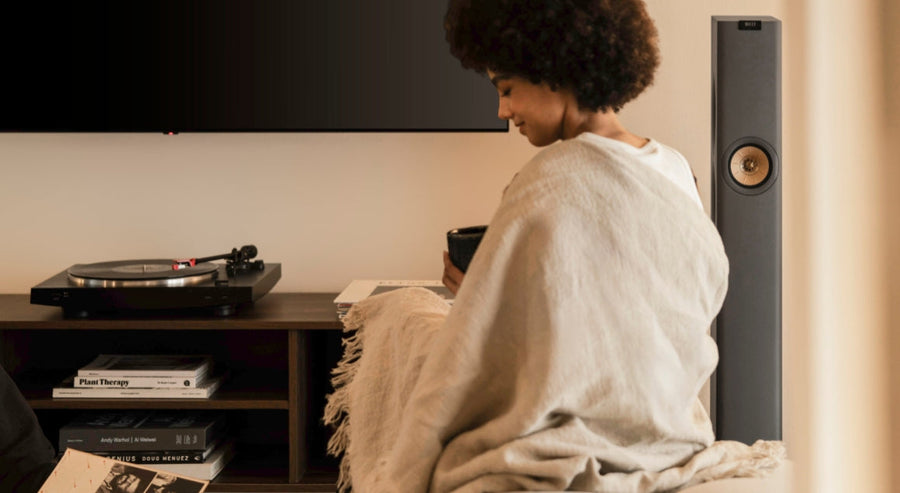 Person relaxing with a cup of coffee in a cosy living room, sitting near a KEF floor-standing speaker and a record player.