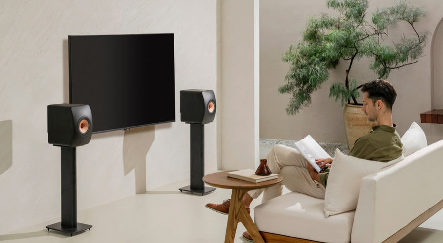 Man relaxing in a modern living room, reading a magazine on a white sofa with KEF stereo speakers on stands flanking a flat-screen TV, showcasing a premium home entertainment setup.
