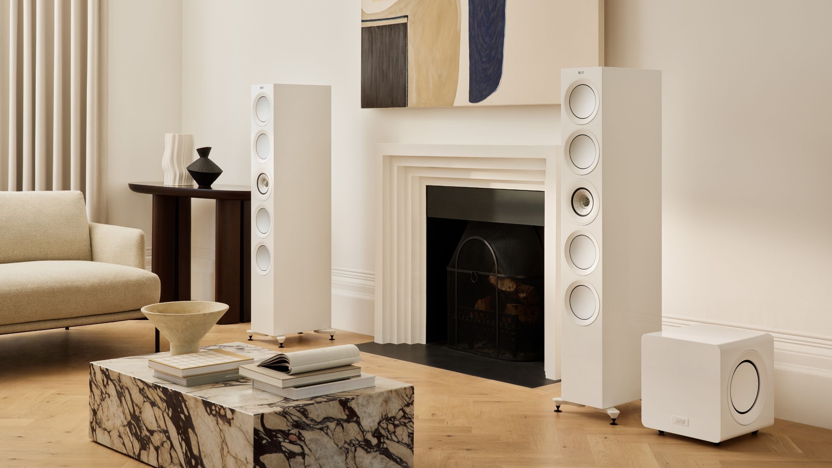 A living room scene showcasing a fireplace, a plush couch, and a white KEF R11 Meta HiFi Speaker positioned nearby with a with KC92 subwoofer