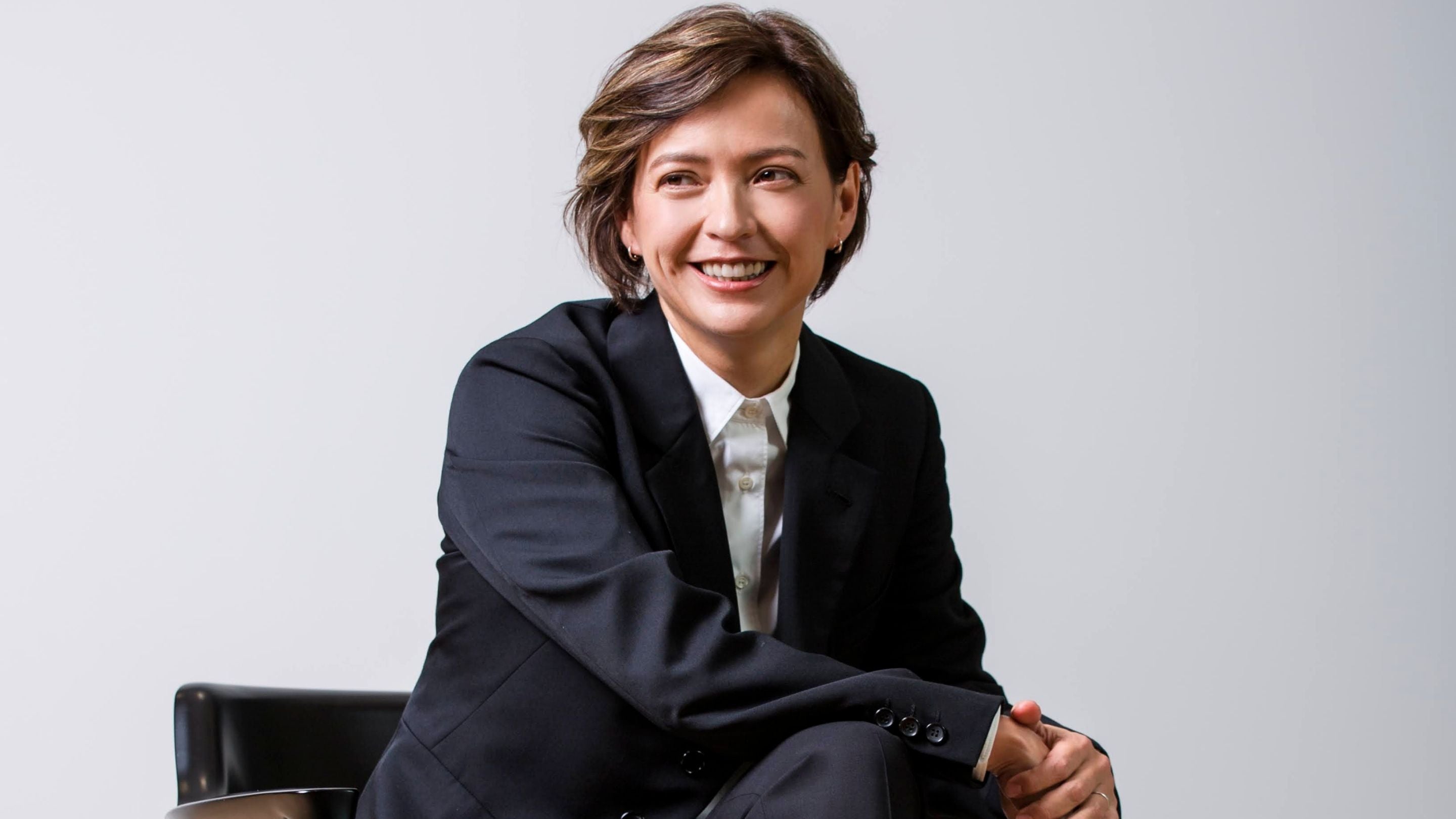 Woman in a black suit sitting on a chair against a grey background