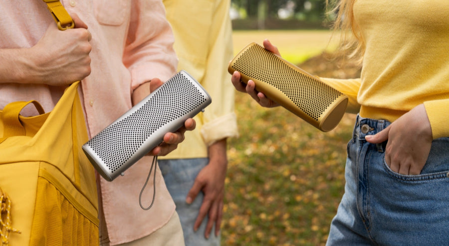 wo people holding portable Bluetooth speakers outdoors, one silver and one gold, during a casual meet-up in the park.