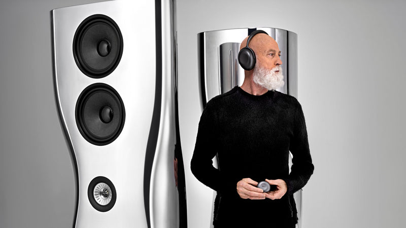 Ross Lovegrove wearing Mu7 headphones and holding Mu3 headphone case in his hand and in the background there is Muon HiFi Speaker, showcasing the products he designed