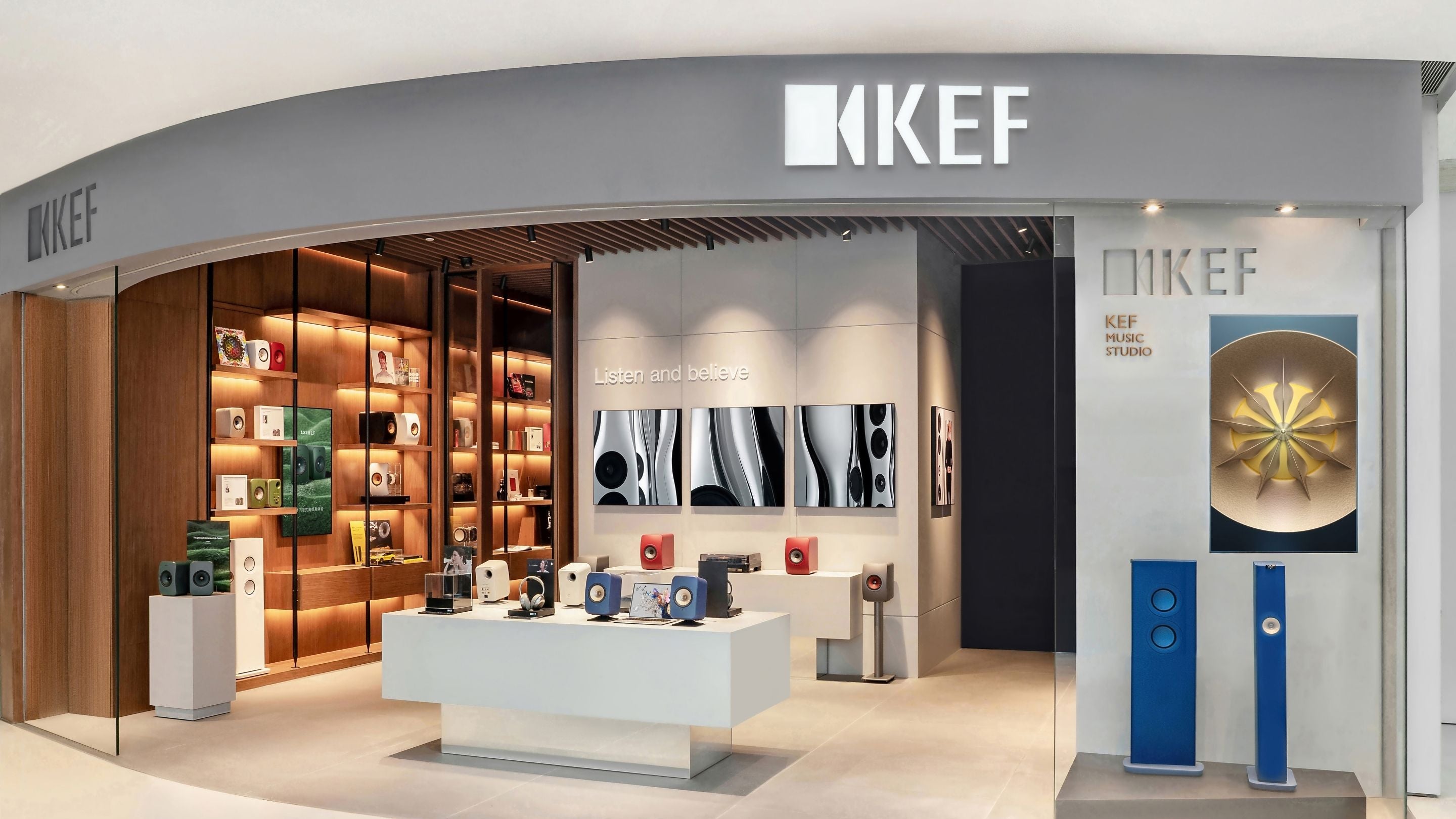 KEF brand retail store interior with a curved gray storefront, illuminated shelving, a central white display table, wall-mounted artwork, and blue speaker units on the right side.