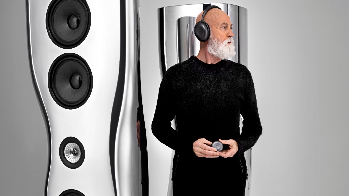 Ross Lovegrove standing in front of large silver Muon floorstanding speakers wearing Mu7 headphones.